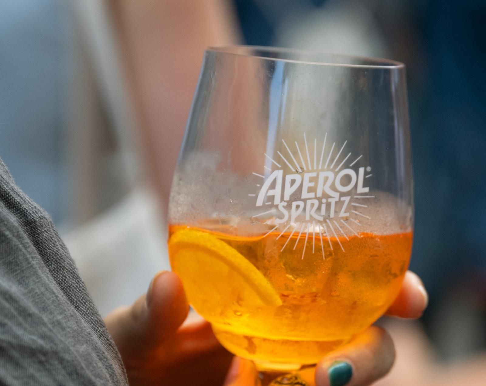 Eine Person hält bei einem geselligen Event ein Glas Aperol Spritz mit einer Orangenscheibe in der Hand.