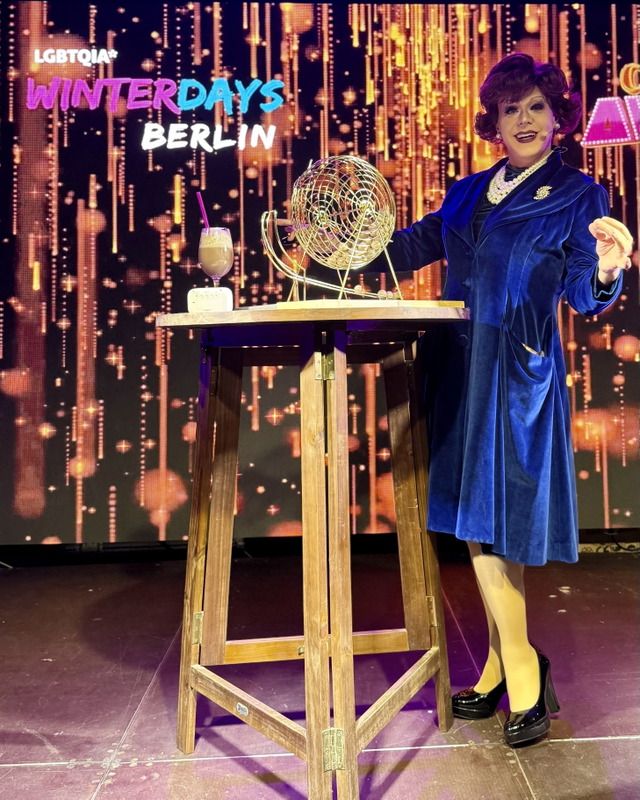 Eine Dragqueen in einem blauen Samtmantel steht auf der Bühne neben einer Tombola und einem Cocktail.