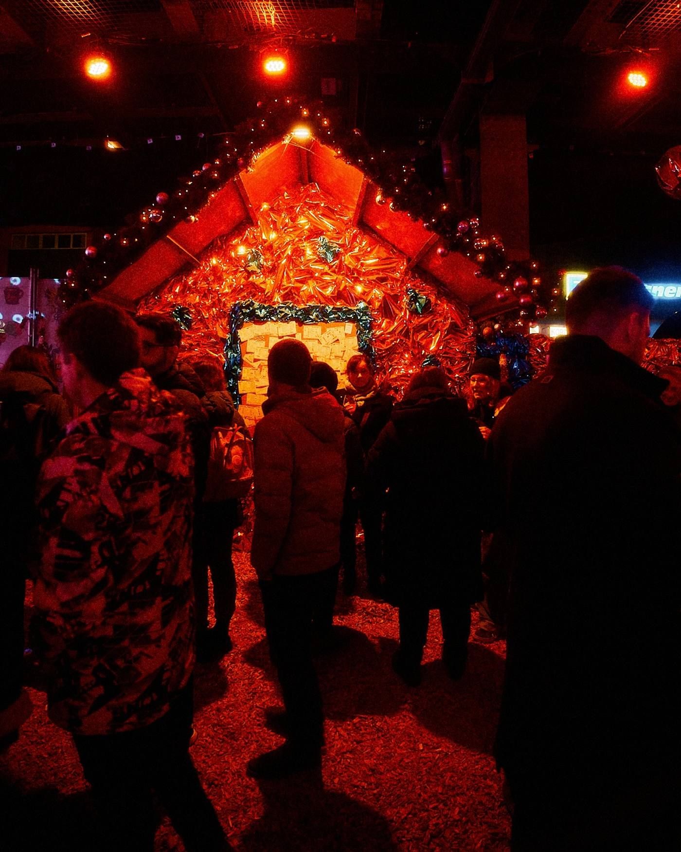 Auf dem Weihnachtsmarkt stehen Besucher vor einer mit Lametta und Lichtern geschmückten, kleinen Hütte im roten Licht.