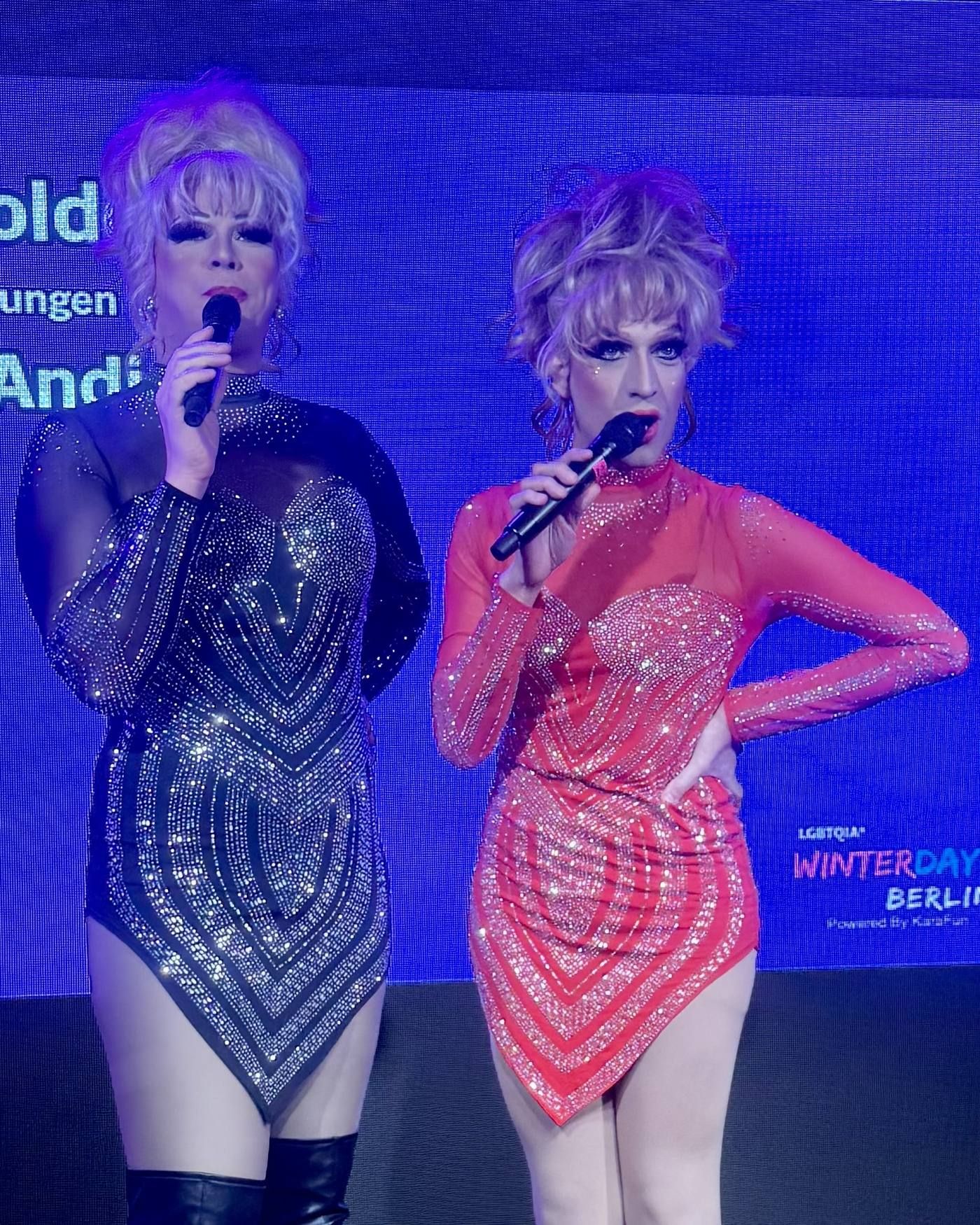 Zwei Drag Queens im Rampenlicht, gekleidet in glitzernde Bühnenoutfits, singen während der LGBTQIA* Winterdays Berlin ins Mikrofon.