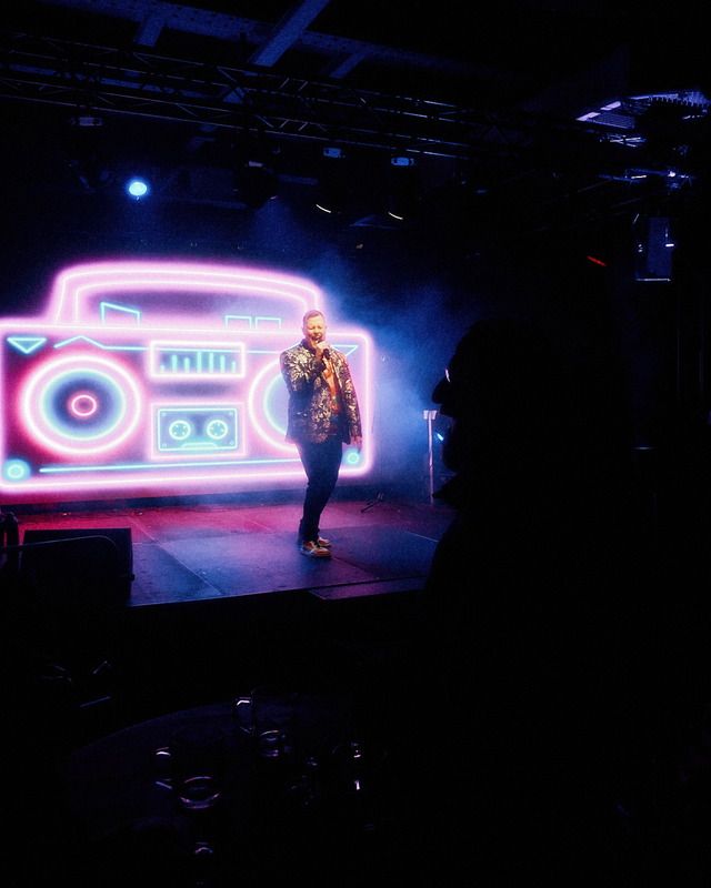 Ein Sänger steht auf einer beleuchteten Bühne vor einer großen, leuchtenden Neon-Boombox und singt in ein Mikrofon.