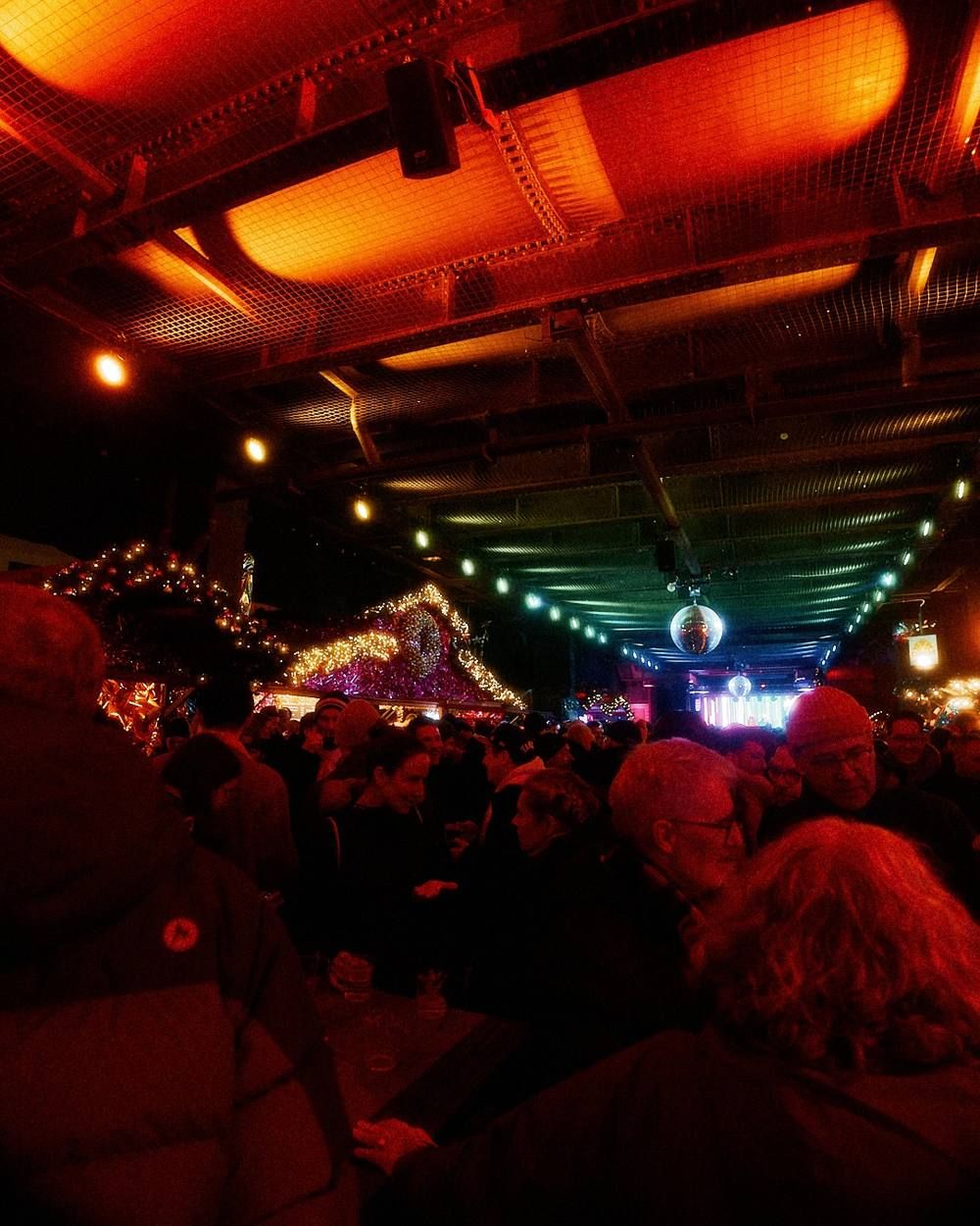 Zahlreiche Besucher unter einer mit Lichterketten geschmückten Überdachung bei einem abendlichen Besuch auf einem weihnachtlichen Markt.