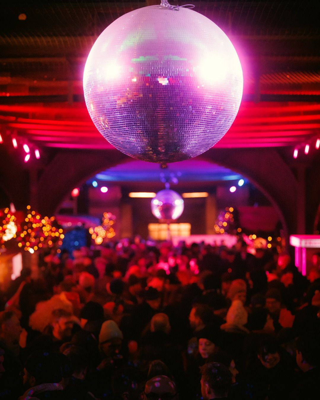 Eine große Disco-Kugel hängt über einer feiernden Menschenmenge, die in rotes Licht getaucht auf einer Party tanzt.