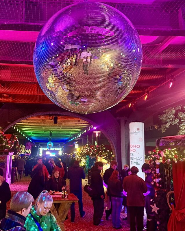 Menschen unter einer riesigen Disco-Kugel auf einem festlich beleuchteten Weihnachtsmarkt mit geschmückten Ständen und bunter Lichtstimmung.
