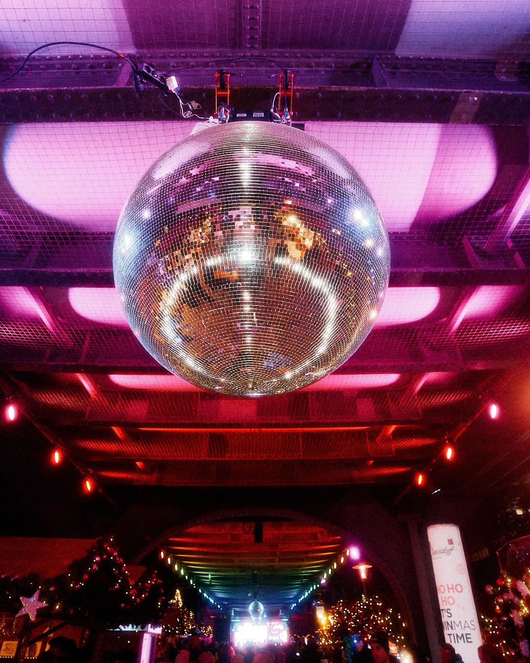 Eine große, glitzernde Disco-Kugel hängt an einer dunklen Brückenkonstruktion, die in atmosphärisches pinkes Licht getaucht ist.