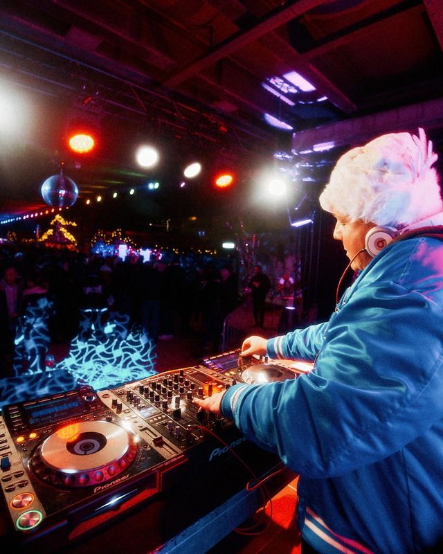 Ein DJ mit weißer Fellmütze legt bei einem nächtlichen Event an einem Pioneer Mischpult vor tanzenden Gästen auf.