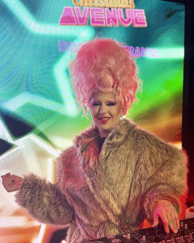 Eine Drag-Künstlerin mit pinker Hochsteckfrisur und Kunstpelzmantel legt fröhlich lächelnd an einem DJ-Pult auf einer Bühne auf.