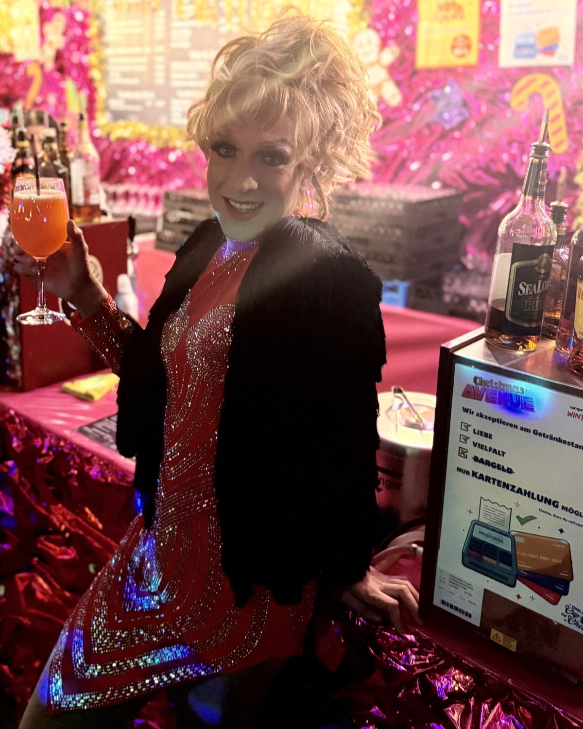 Eine festlich gekleidete Drag-Künstlerin in einem roten Paillettenkleid hält ein Glas mit einem Cocktail an einem Weihnachtsmarktstand.