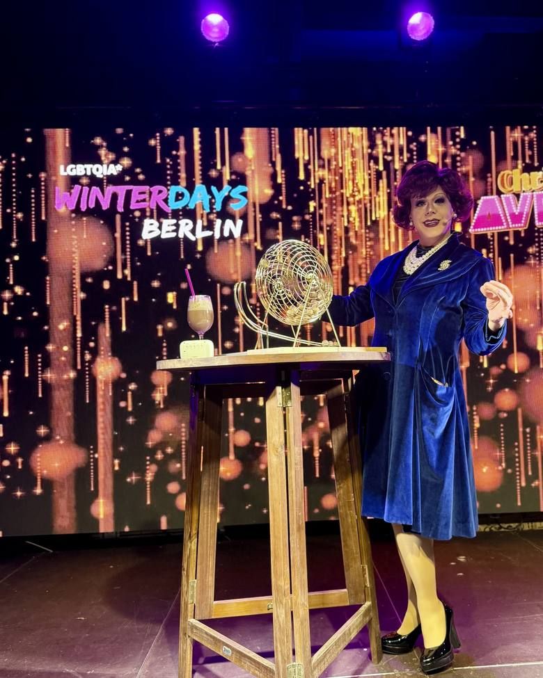 Eine elegant gekleidete Drag-Moderatorin präsentiert eine Bingotrommel auf der Bühne des LGBTQIA Winterdays Berlin Events.