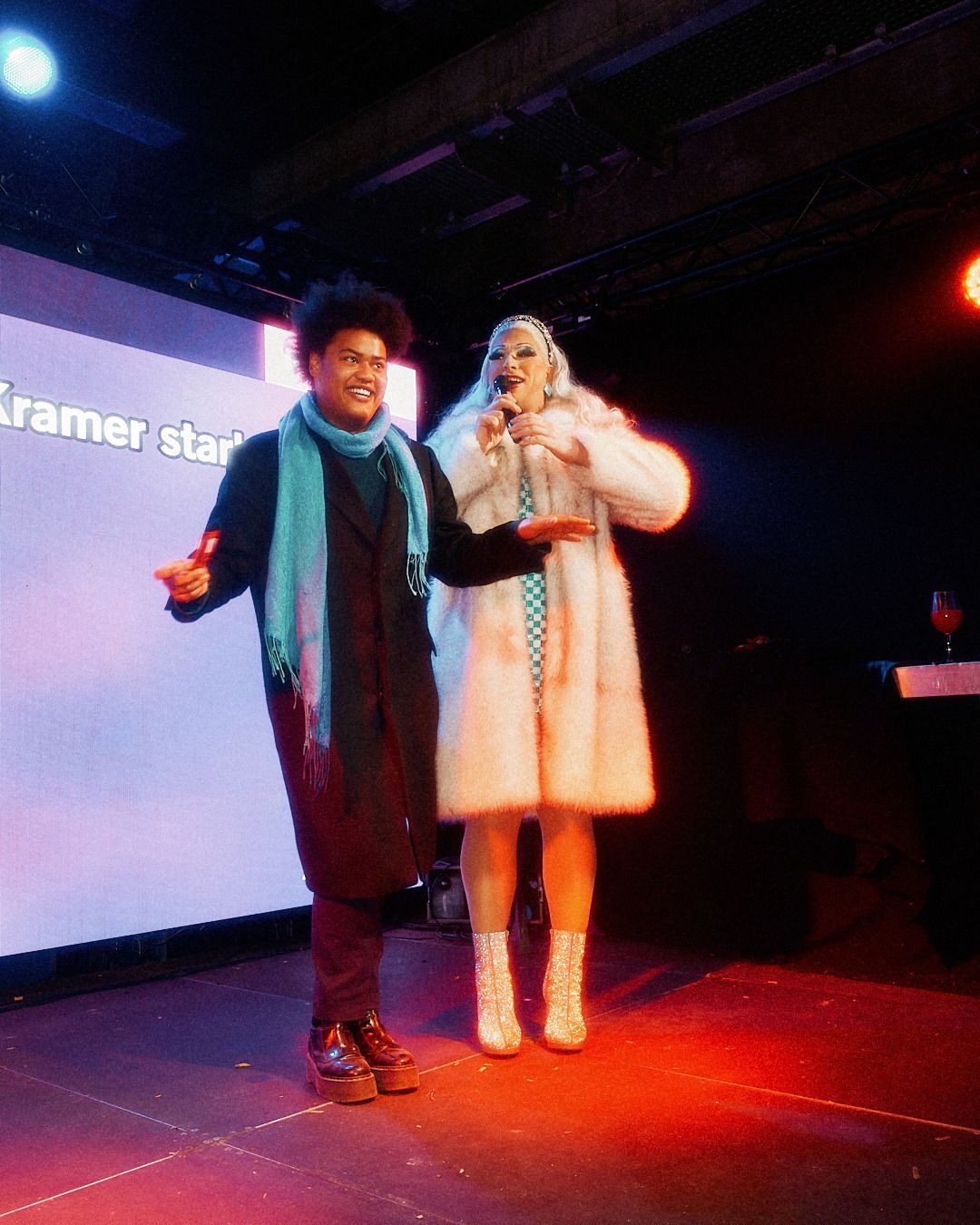 Eine Dragqueen und ein Moderator stehen auf einer beleuchteten Bühne und führen gemeinsam durch das Eventprogramm.