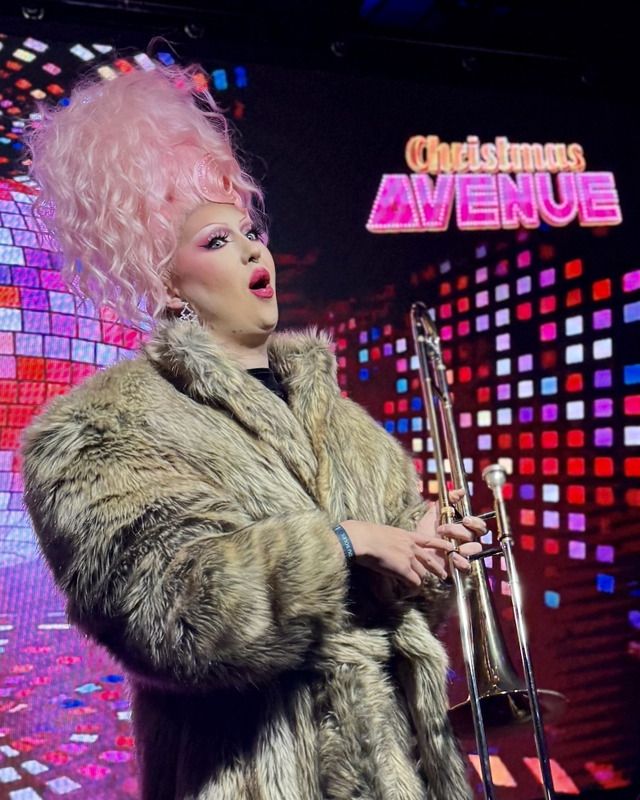 Eine Dragqueen mit pinker Hochsteckfrisur und Pelzmantel spielt auf einer Bühne eine Posaune vor einem bunten Hintergrund.