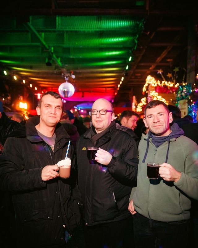 Drei Männer stehen auf einem beleuchteten Weihnachtsmarkt und halten heiße Getränke in Glasbechern in die Kamera.