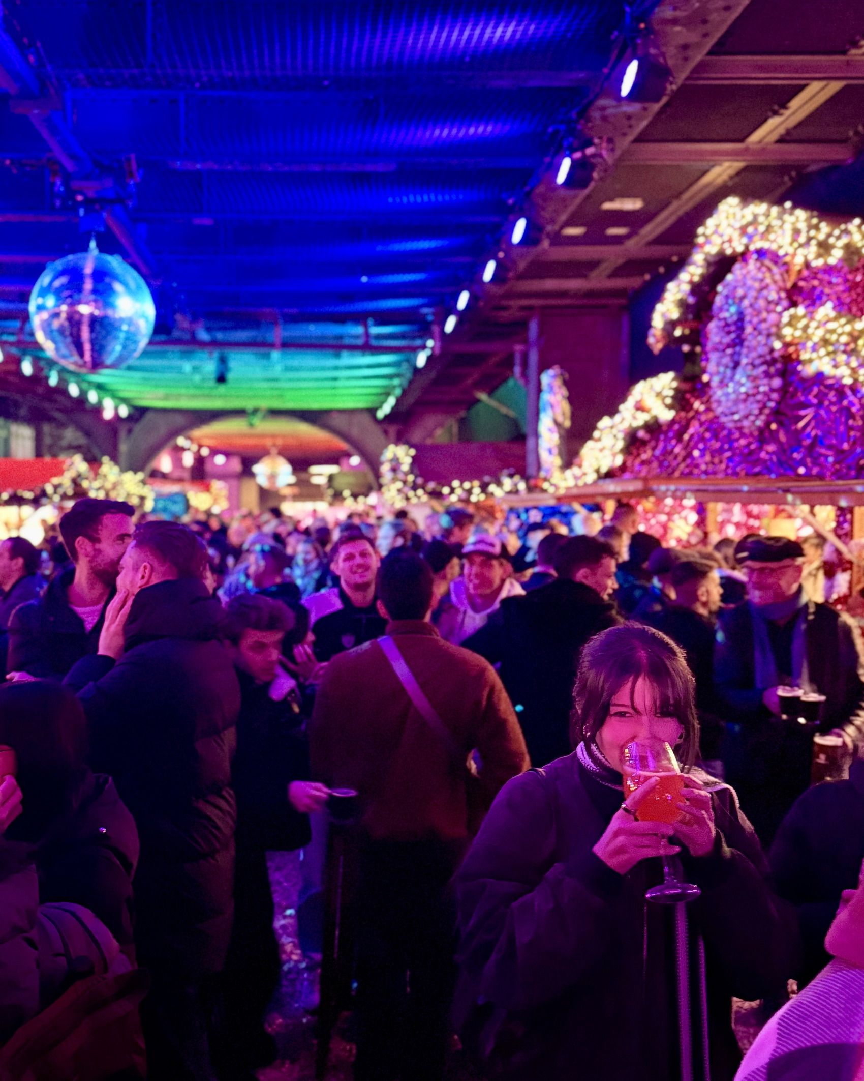 Eine junge Frau trinkt lächelnd einen Cocktail in einer belebten, festlich beleuchteten Eventlocation mit einer Discokugel.