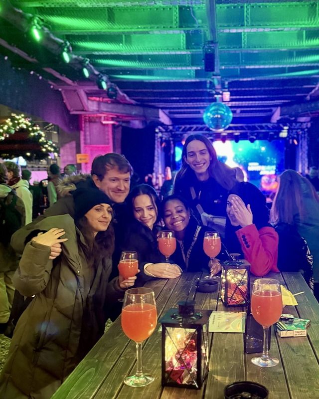 Eine Gruppe von sechs jungen Erwachsenen sitzt an einem Holztisch, lacht gemeinsam und stößt mit Cocktails an.