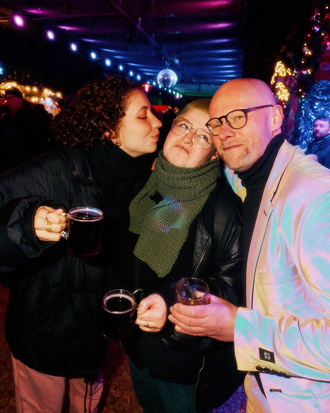 Drei Freunde stehen bei festlicher Beleuchtung zusammen, halten Glühweintassen in der Hand und genießen gemeinsam den Abend.
