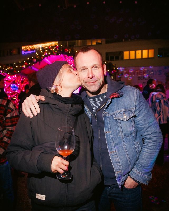Eine Frau mit Mütze küsst einen Mann auf die Wange, während sie ein Glas Glühwein auf einem Weihnachtsmarkt hält.