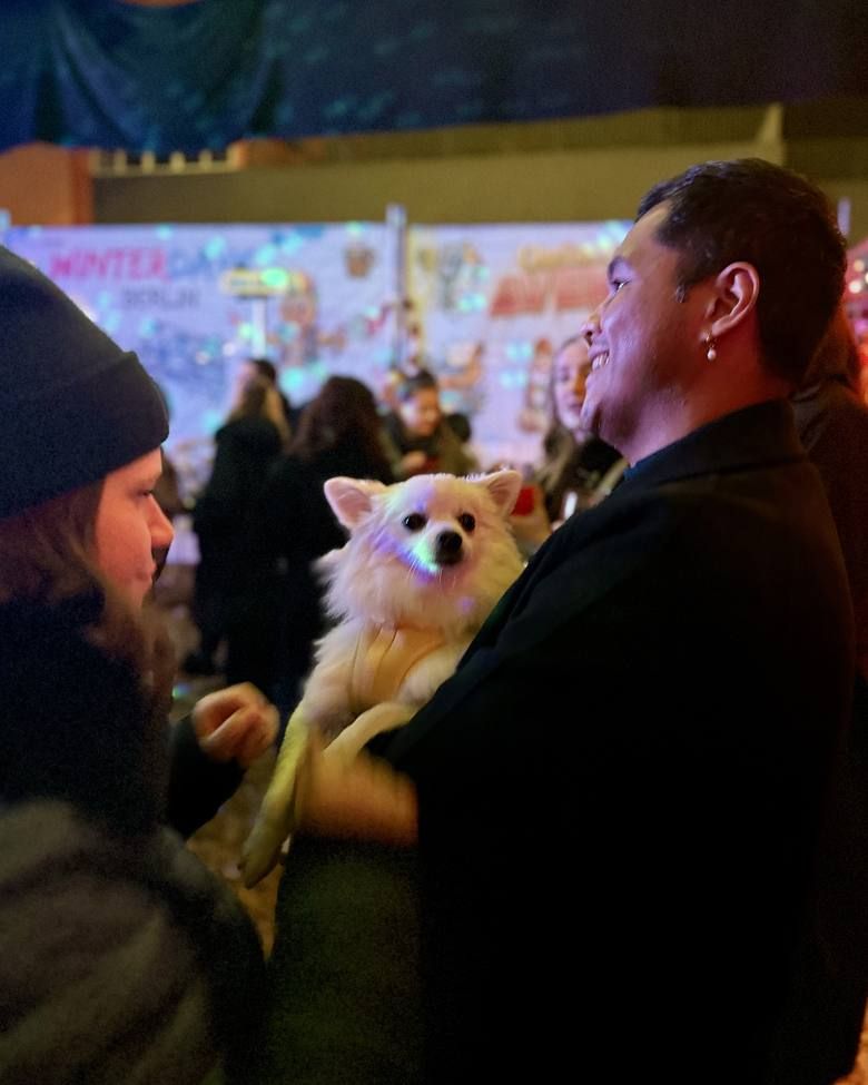 Ein lächelnder Mann hält einen kleinen weißen Hund im Arm, während er sich mit einer Person unterhält.