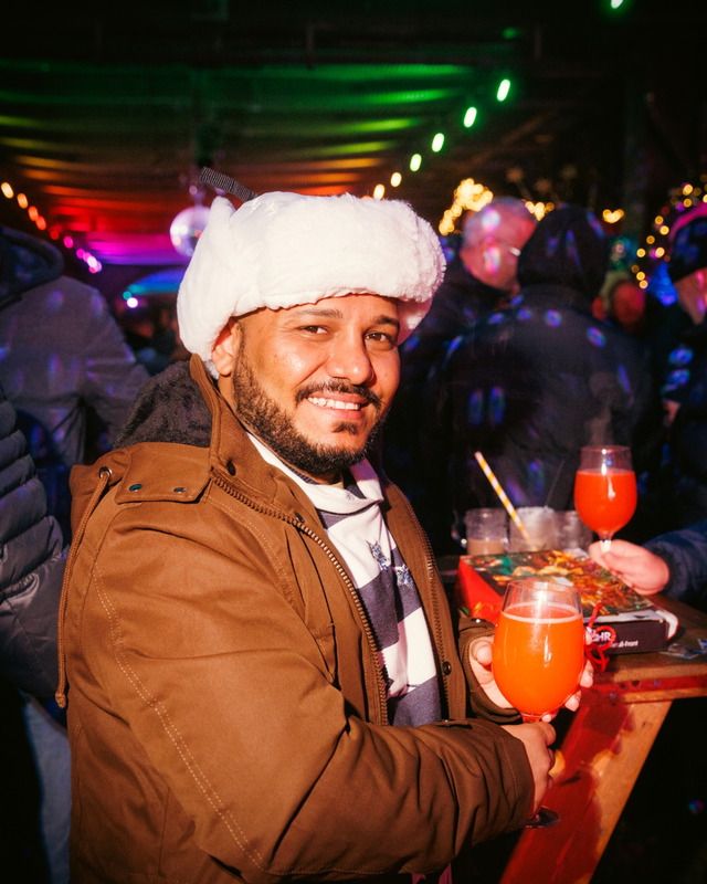 Ein lächelnder Mann mit weißer Pelzmütze hält ein orangefarbenes Getränk in einer festlich beleuchteten, abendlichen Umgebung in die Kamera.