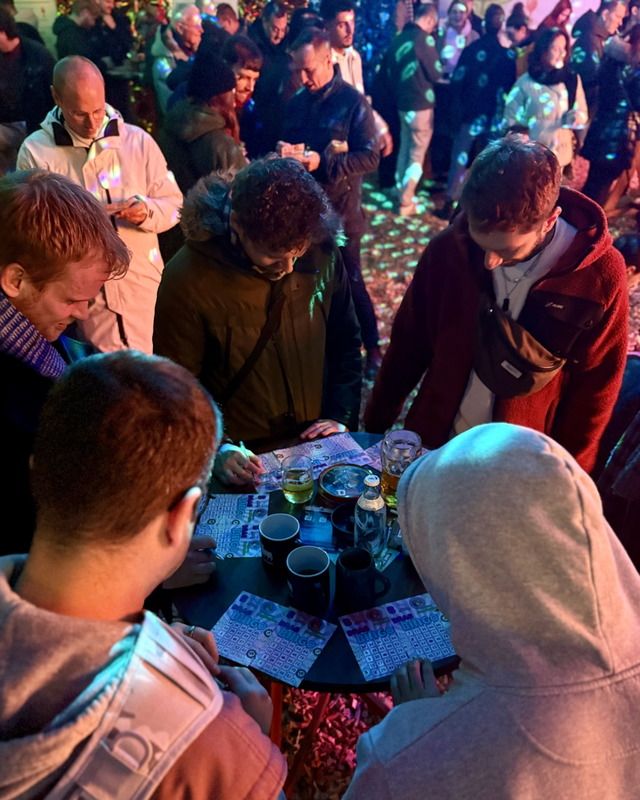 Eine Gruppe junger Leute spielt konzentriert Bingo an einem runden Tisch in einer stimmungsvoll beleuchteten, feiernden Menge.