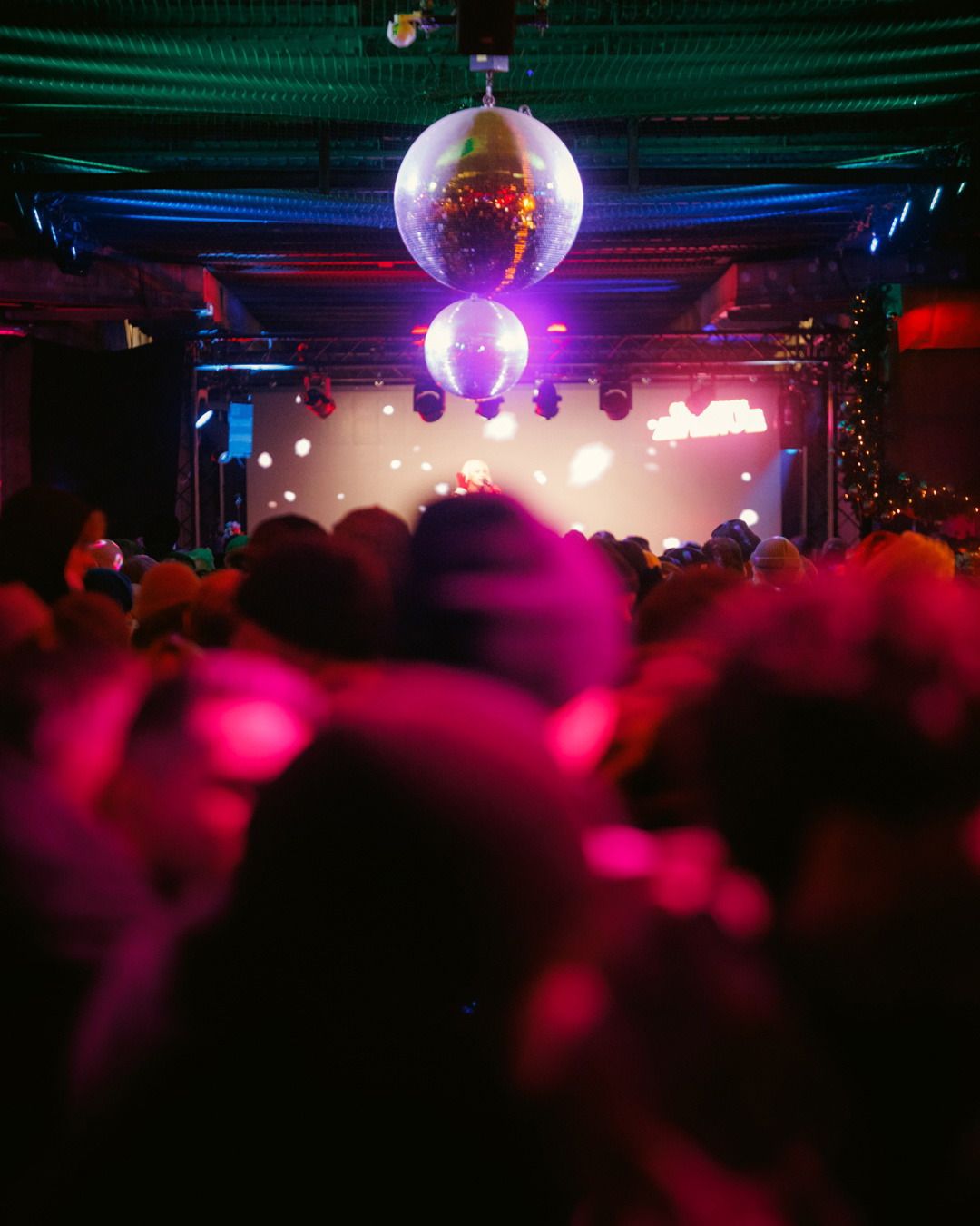 Eine dicht gedrängte Menschenmenge feiert ausgelassen vor einer beleuchteten Bühne unter zwei hängenden, glitzernden Discokugeln in einer Eventhalle.