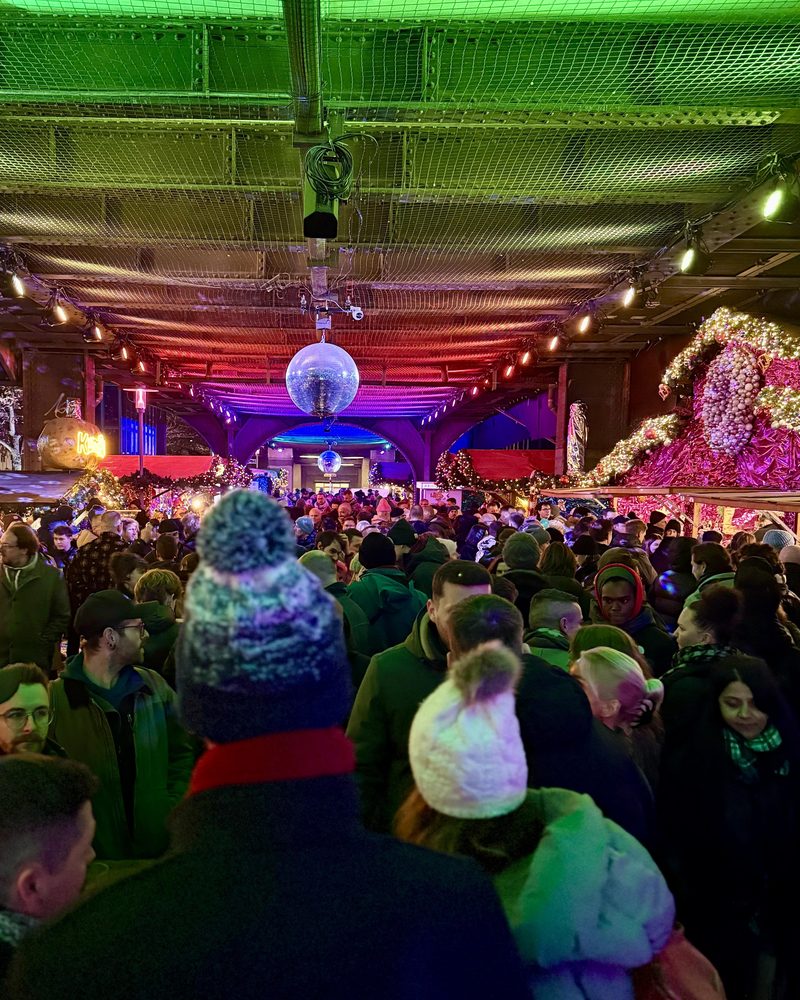 Zahlreiche Besucher drängen sich unter festlich beleuchteten Brückenbögen mit hängenden Discokugeln auf einem dicht gefüllten abendlichen Weihnachtsmarkt.