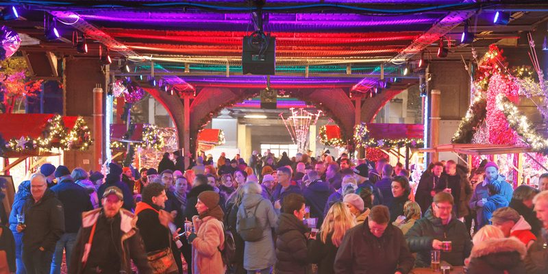 Zahlreiche Besucher unter bunter Beleuchtung feiern und unterhalten sich gesellig auf einem festlich geschmückten Weihnachtsmarkt mit Marktständen.