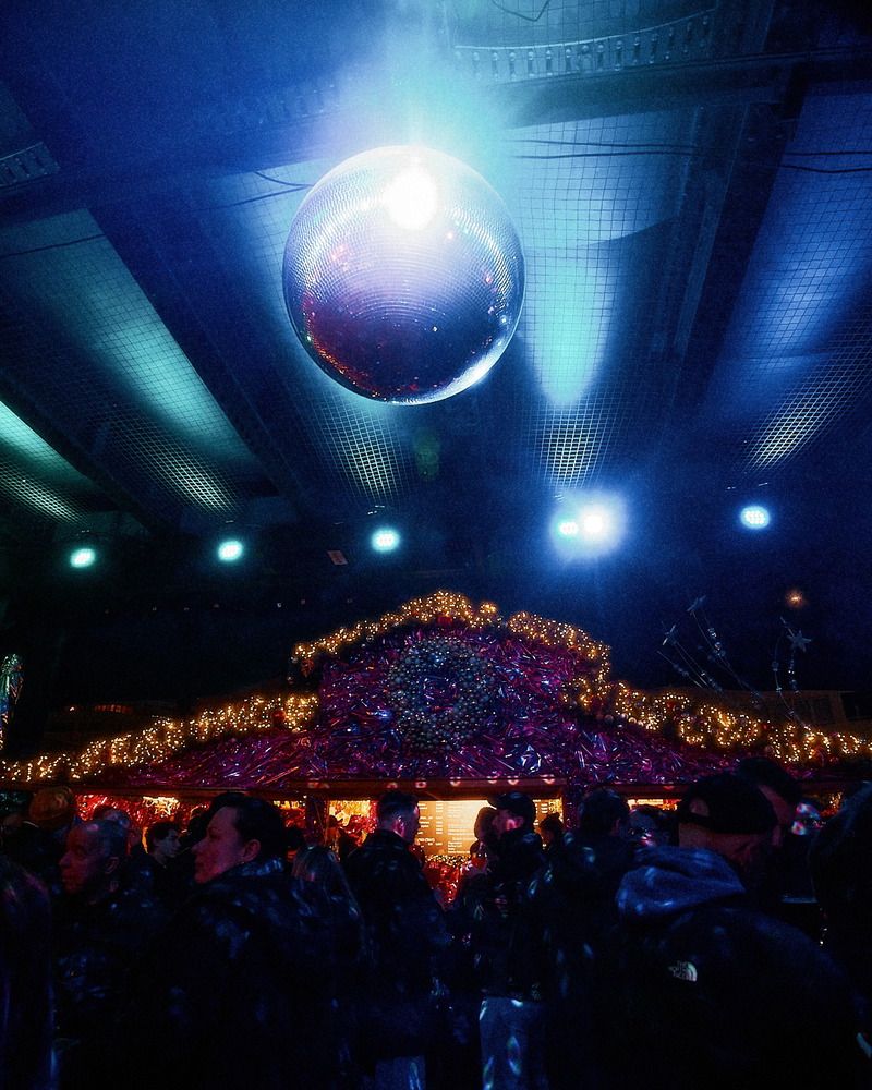 Eine funkelnde Disco-Kugel hängt über einem festlich beleuchteten Stand, während zahlreiche Besucher abends auf dem Weihnachtsmarkt feiern.
