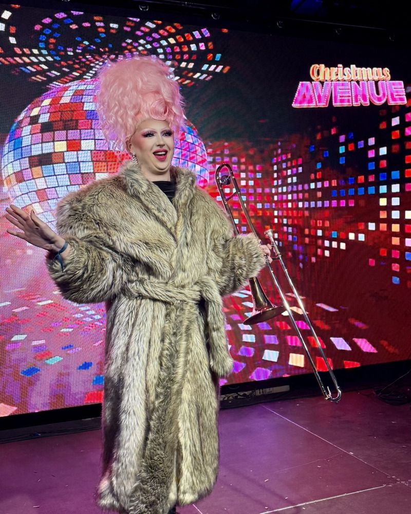 Eine Drag-Künstlerin mit pinker Hochsteckfrisur und Pelzmantel hält eine Posaune vor einer leuchtenden Disco-Grafik auf der Bühne.