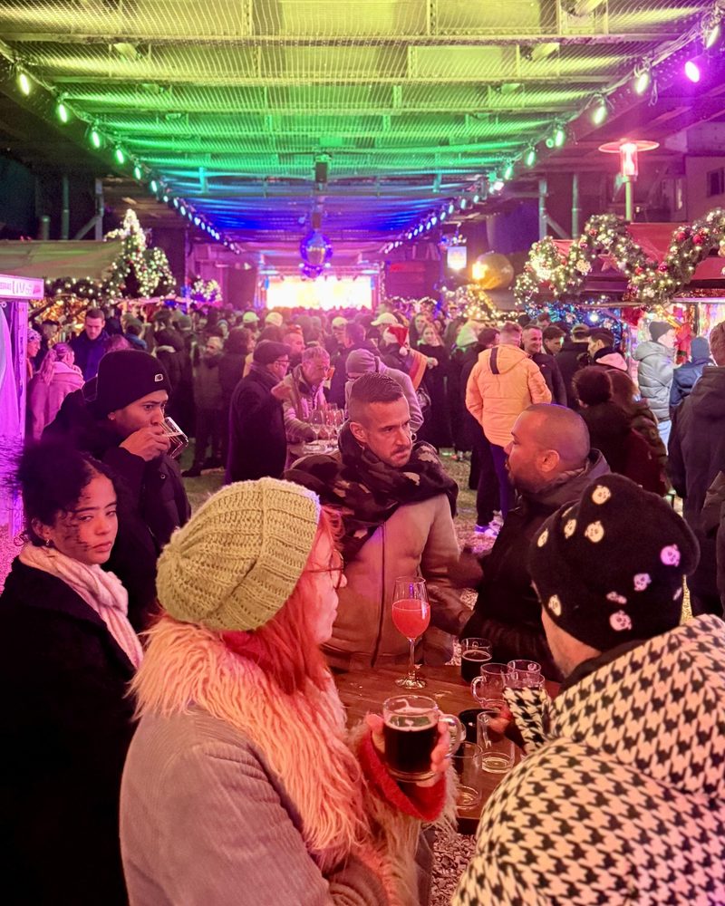 Eine Gruppe von Gästen trinkt an einem Stehtisch auf einem beleuchteten Weihnachtsmarkt und unterhält sich lebhaft miteinander.