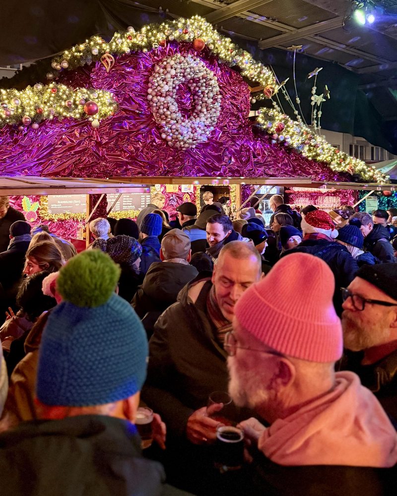 Zahlreiche Besucher stehen eng beieinander an einem festlich geschmückten Weihnachtsmarktstand und trinken gemeinsam in geselliger Atmosphäre.
