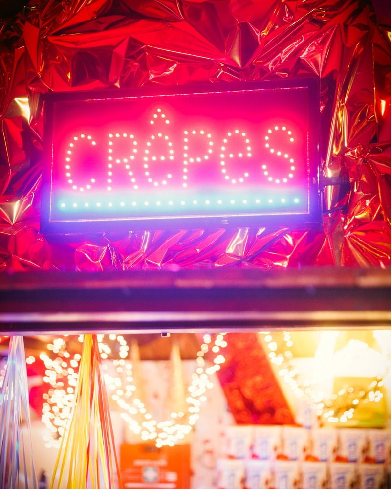 Ein leuchtendes Neon-Schild mit der Aufschrift Crêpes hängt vor einer glänzenden, roten Folie auf einem festlichen Weihnachtsmarktstand.
