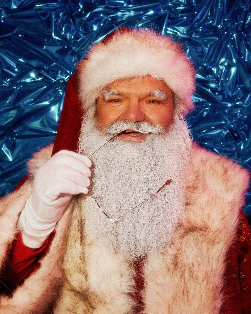 Ein freundlicher Weihnachtsmann mit weißem Bart hält seine Brille an den Lippen vor einem glitzernden blauen Hintergrund.