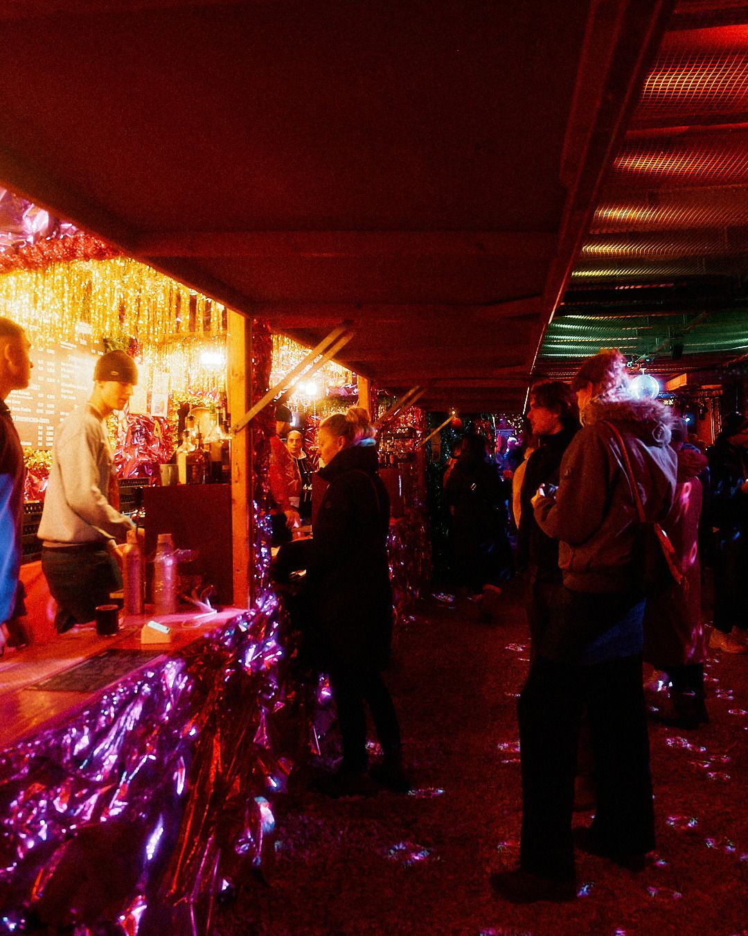 Gäste stehen an einem festlich beleuchteten Tresen, während ein Verkäufer Getränke in einer festlichen, pink glitzernden Umgebung ausschenkt.
