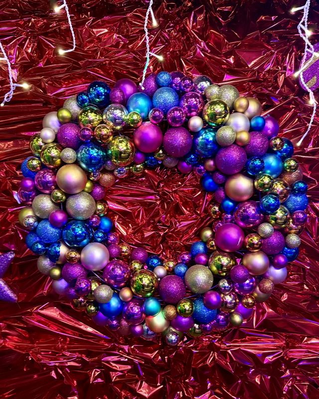 Ein farbenfroher Weihnachtskranz aus glitzernden blauen, violetten und goldenen Christbaumkugeln hängt vor einem knittrigen, reflektierenden roten Hintergrund.