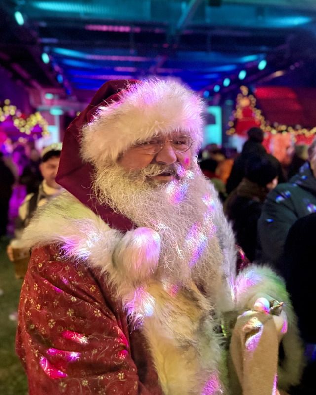 Ein freundlicher Weihnachtsmann mit langem weißen Bart lächelt in einer festlich beleuchteten Umgebung mit bunten Lichtreflexen.