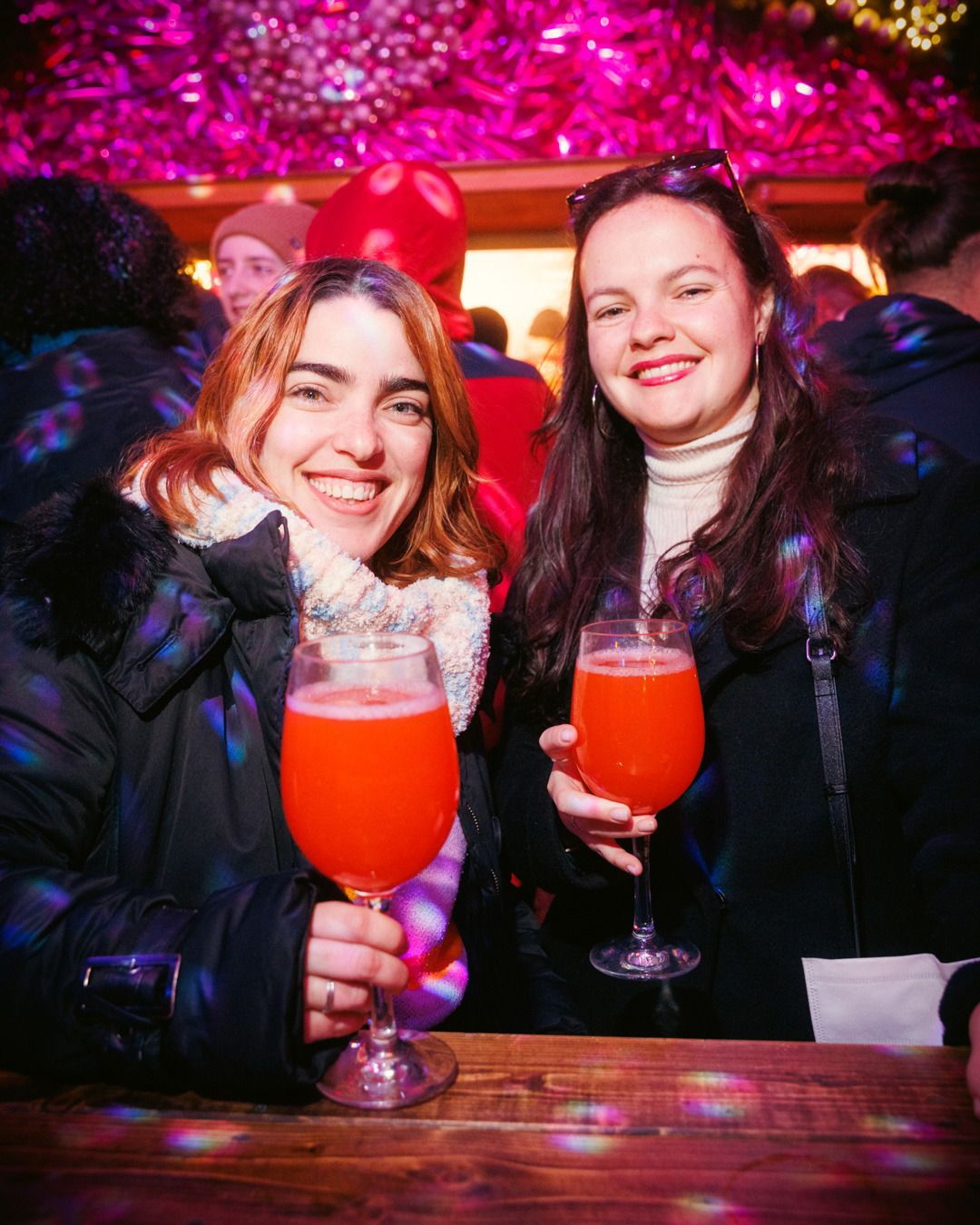 Zwei lächelnde Frauen halten bei festlicher Beleuchtung orangefarbene Cocktails in den Händen und blicken in die Kamera.