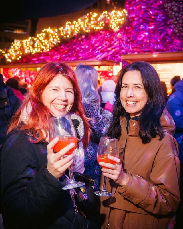 Zwei lachende Frauen halten Weingläser mit einem orangefarbenen Getränk in der Hand und feiern auf einem Weihnachtsmarkt.