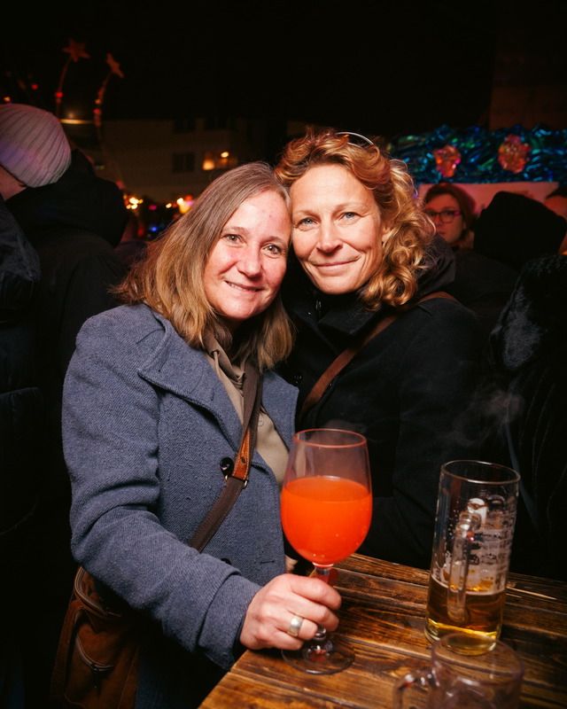 Zwei freundlich lächelnde Frauen stehen eng beieinander an einem Holztisch und halten ein Glas mit einem orangen Getränk.