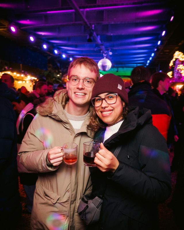 Zwei glückliche Gäste stoßen auf einem abendlich beleuchteten Weihnachtsmarkt mit heißen Getränken in Glasbechern gemeinsam an.