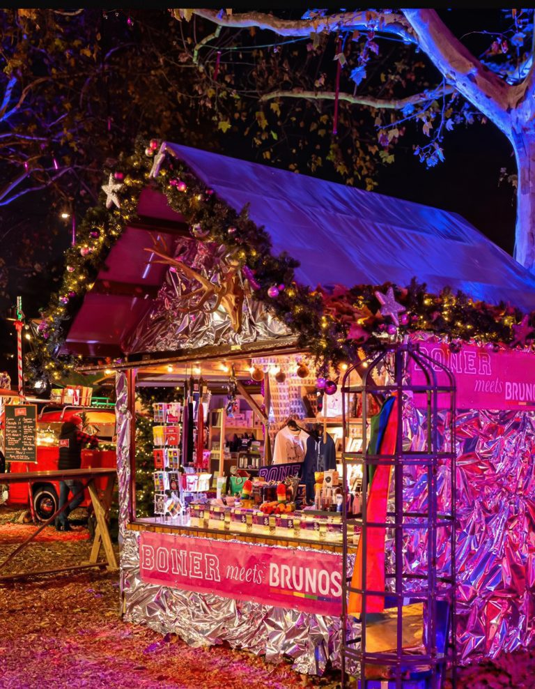 Festlich geschmückter Stand auf der Christmas Avenue Berlin mit dem Schriftzug ‚BONER meets BRUNOS‘, beleuchtet von bunten Lichtern und umgeben von weihnachtlicher Dekoration