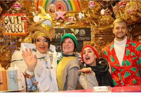 „Gaumerausch auf der Christmas Avenue – Bunte Vielfalt auf Berlins LGBTQIA* Weihnachtsmarkt“ Fröhliche Standbetreiber auf dem LGBTQIA* Weihnachtsmarkt Christmas Avenue in Berlin, verkleidet in bunten Kostümen am Gaumerausch-Stand