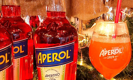 Neben einem mit einer Orangenscheibe garnierten Weinglas glänzen Aperol-Flaschen und fangen die lebendige Essenz der Berliner Happy Hour ein. Das Ambiente ist hell und festlich, und Lichtreflexe unterstreichen die Ästhetik.